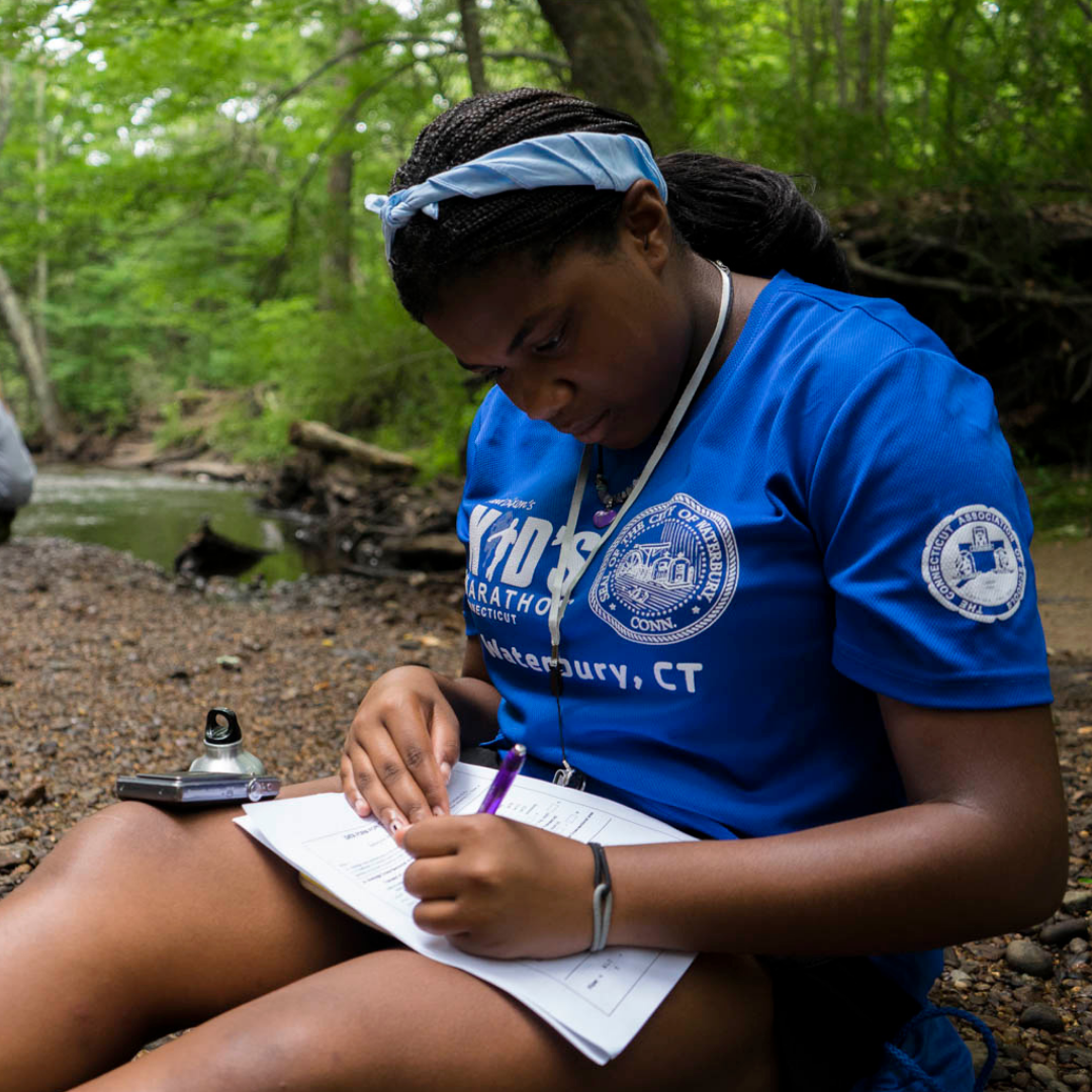 UConn student sitting in woods writing in notebook